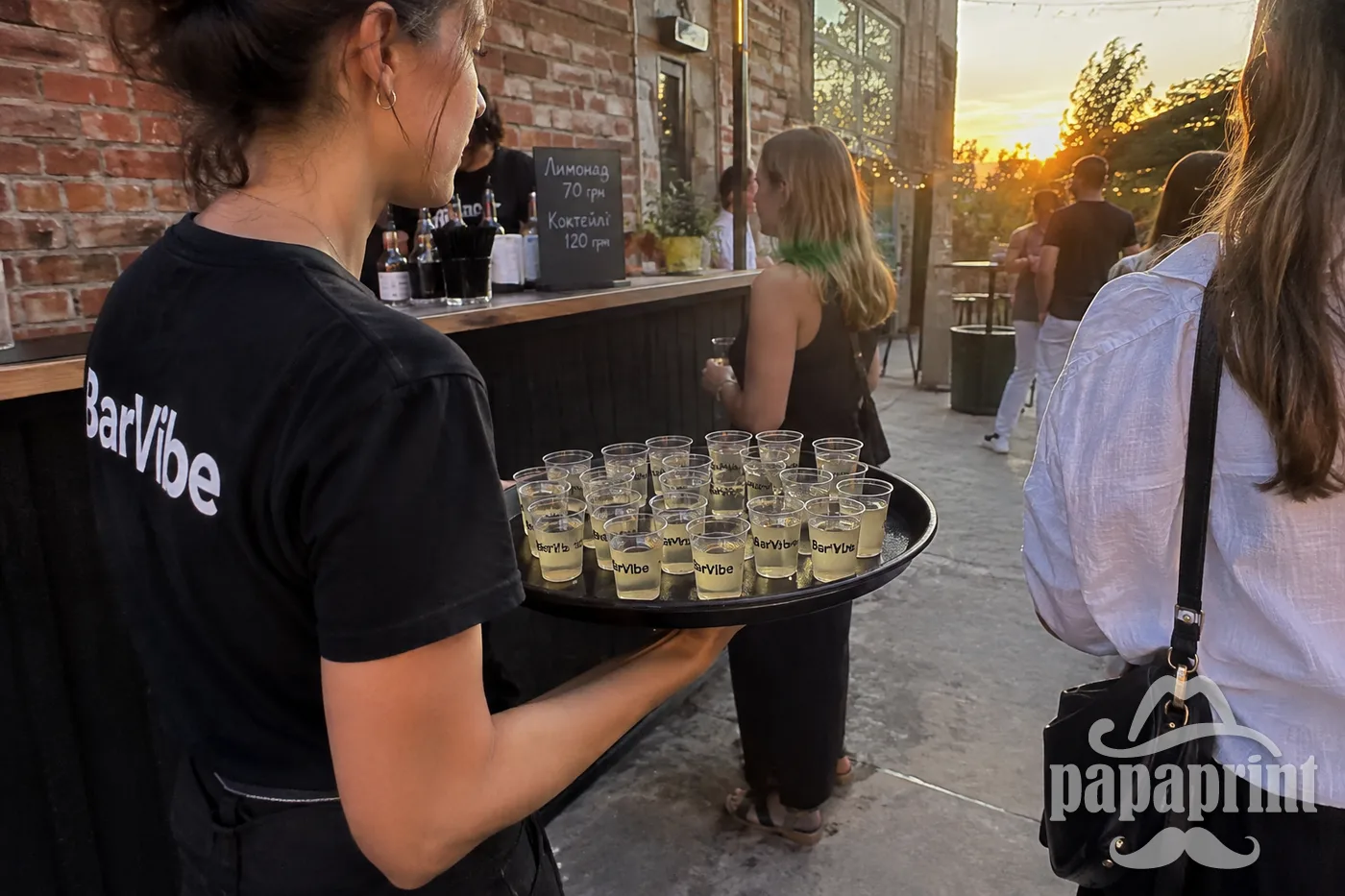 Офіціант несе піднос із прозорими шот-чарками BarVibe у дворі біля цегляної стіни.