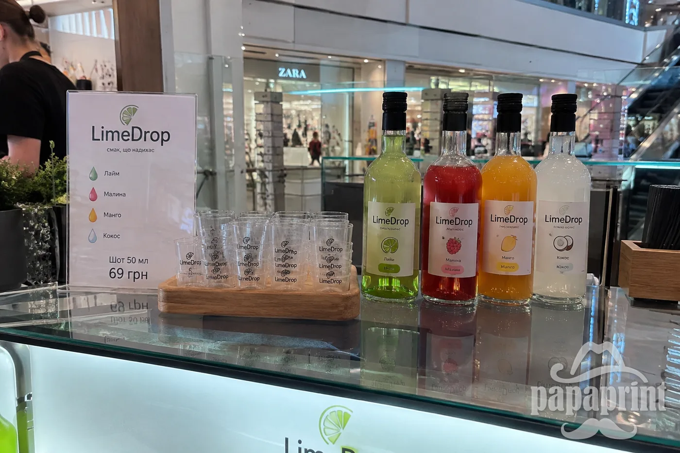 Прозорі шот-чарки LimeDrop з простим логотипом стоять біля пляшок у кіоску ТРЦ.
