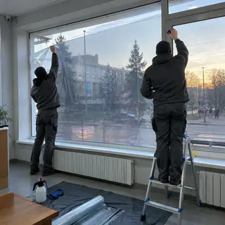 Двоє монтажників клеять товсту плівку на вітрину першого поверху зсередини (ілюстрація)
